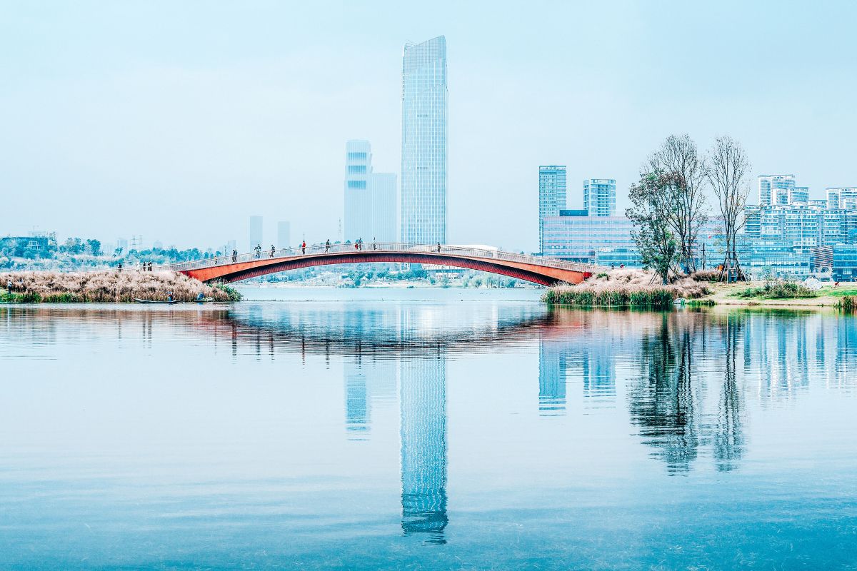 Scenic view of a red pedestrian bridge over a calm lake with modern buildings in the background in Chengdu, China.