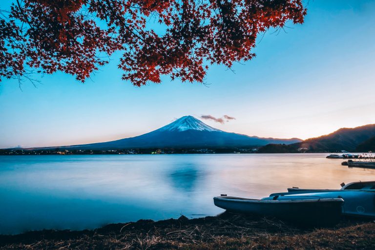 The BEST 2-day Itinerary in Mount Fuji: Snow-capped Mount Fuji viewed across a calm lake at sunset, with boats on the shore and red maple leaves framing the scene.