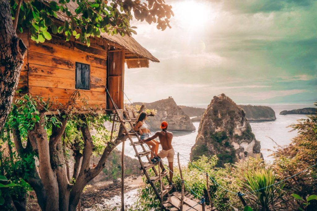 Best Hotels in Bali for Couples (Romantic Getaways 2025): Couple enjoying a romantic moment on a wooden treehouse with a scenic ocean view and rocky cliffs in Nusa Penida, Bali at sunset.