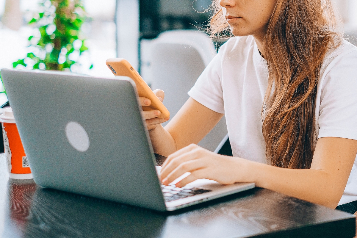 A digital nomad working on a laptop while checking a phone in a cafe, using her Best Digital Nomad Essentials for remote work and travel.