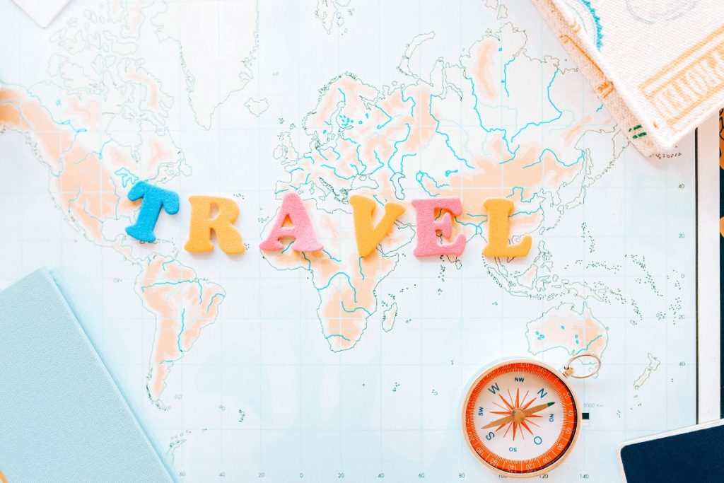 Flat lay of a world map with colourful letters spelling “TRAVEL” and a compass, symbolising adventure and the Best Travel Insurance for Travellers in 2025.