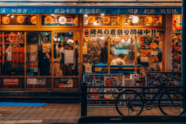 Japanese restaurant in Tokyo with colorful posters, hanging lanterns, and people dining inside, captured at night for a Tokyo Food Guide.