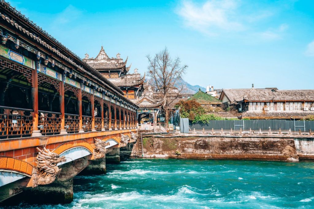 Traditional Chinese bridge decorated with dragon carvings crossing over turquoise river water, with historic wooden buildings and temples in the background on a clear day in Chengdu, perfect for a travel guide on where to stay in Chengdu.