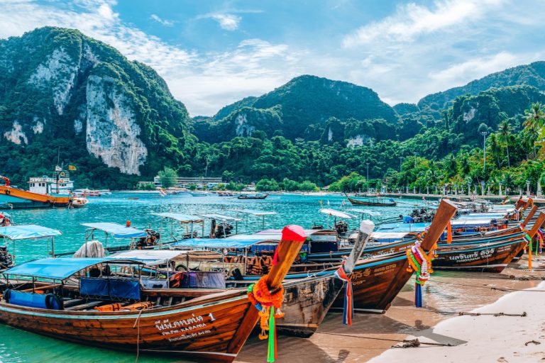 Traditional wooden longtail boats decorated with colorful ribbons line the sandy shore in front of turquoise waters and lush green cliffs, a scenic view often found when exploring where to stay in Phuket.