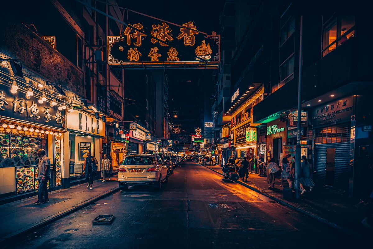 A vibrant Hong Kong street at night glowing with neon restaurant signs and warm yellow lights. People walk past food stalls and eateries as cars line the narrow road, creating a lively atmosphere that captures The BEST Food Spots in Hong Kong.