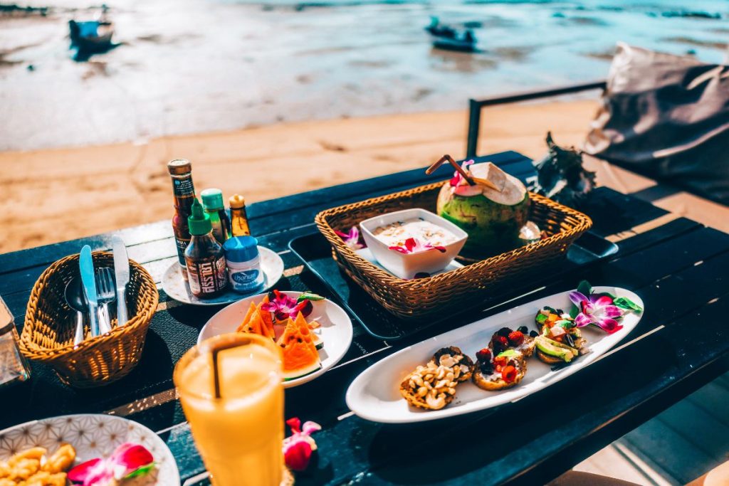 A colorful beachfront brunch spread in Phuket featuring tropical dishes like fruit slices, avocado toasts, and a fresh coconut drink served on a dark wooden table overlooking the sea. Perfect visual for highlighting top food spots in Phuket.