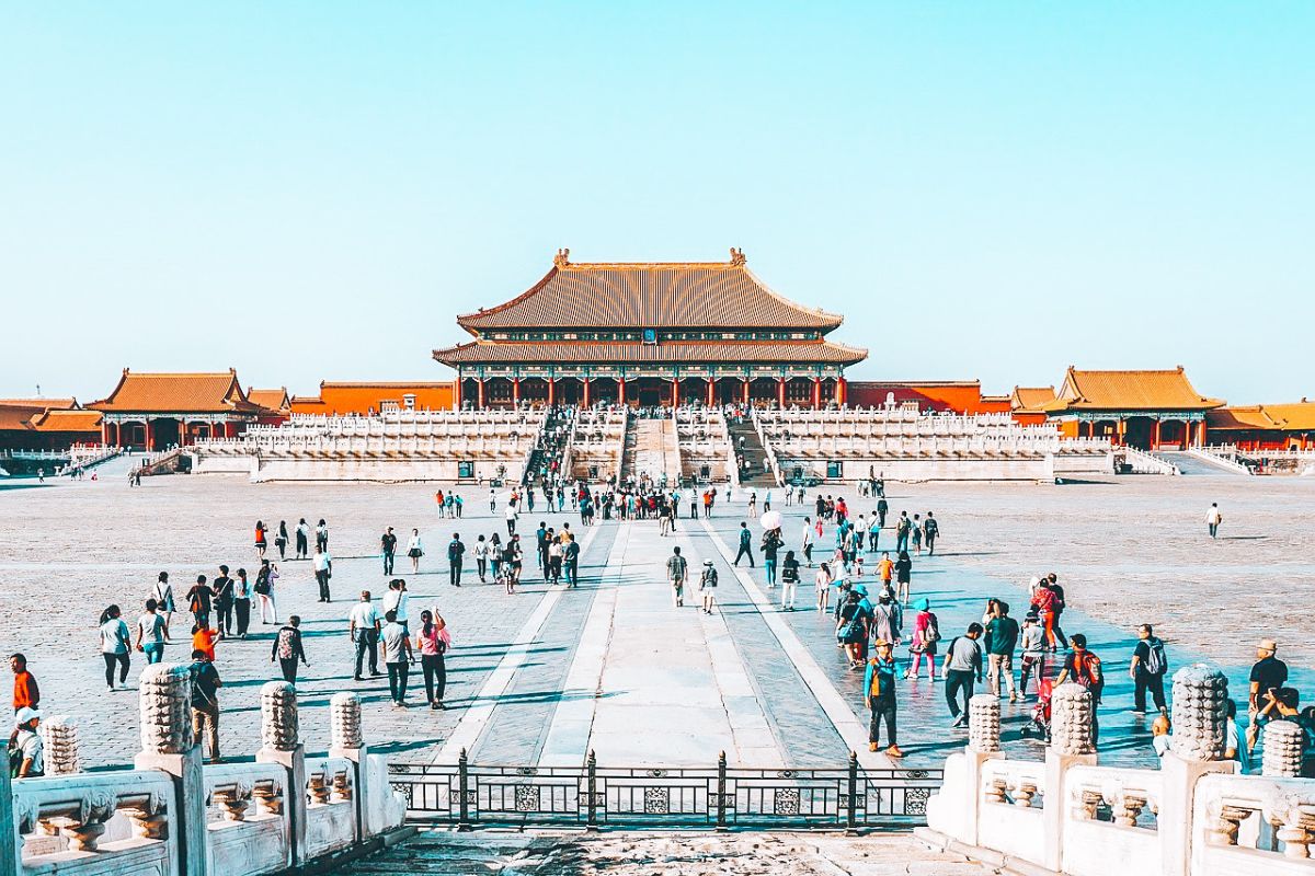 Visitors walking across a wide stone courtyard toward the ornate golden-roofed palace in the Forbidden City under a clear blue sky. This iconic landmark scene complements travel guides about Food Spots in Shanghai by showcasing nearby cultural attractions in China.