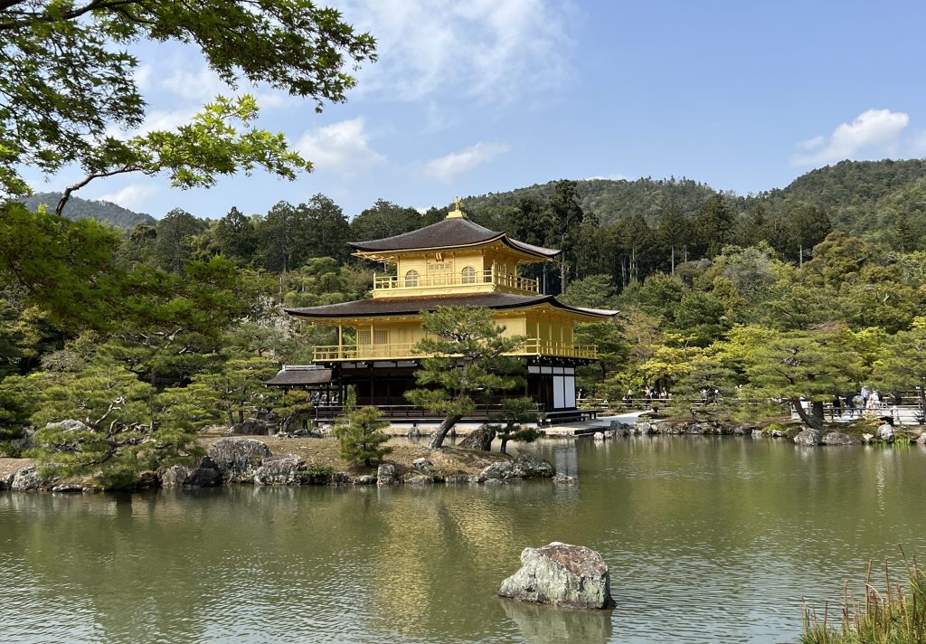 A traditional Shinto temple decorated in gold leaf sits atop a reflective pond, surrounded by rocks and trees.
